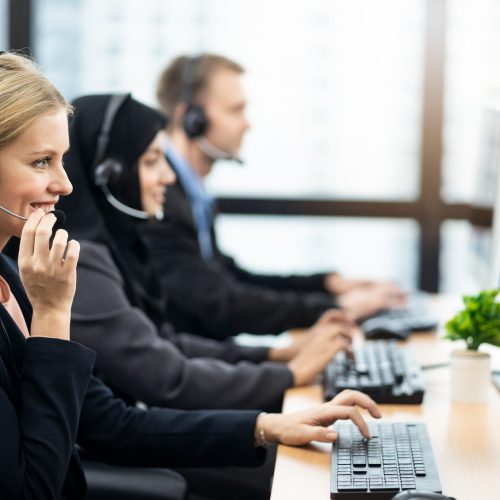 Beautiful Caucasian female call center with headset talking to customer in office. The girl holding headphone microphone with smile. Seen from woman side with colleagues muslim girl and Caucasian man.
