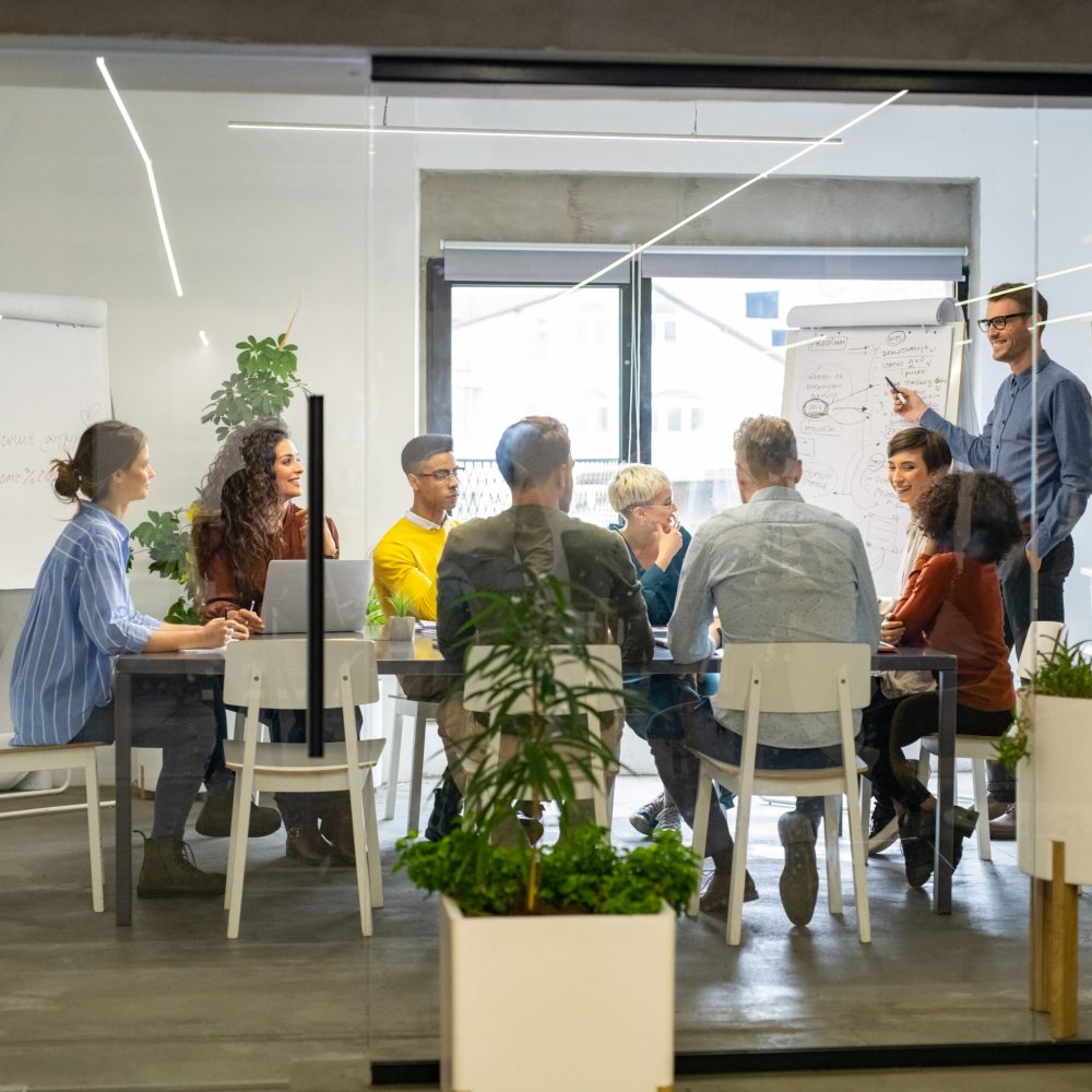 Confident mature businessman giving a presentation to his crative team in office. Business brief with annual goals with casual employees. Happy leadership man training young businessmen and smiling businesswomen sitting at conference table.